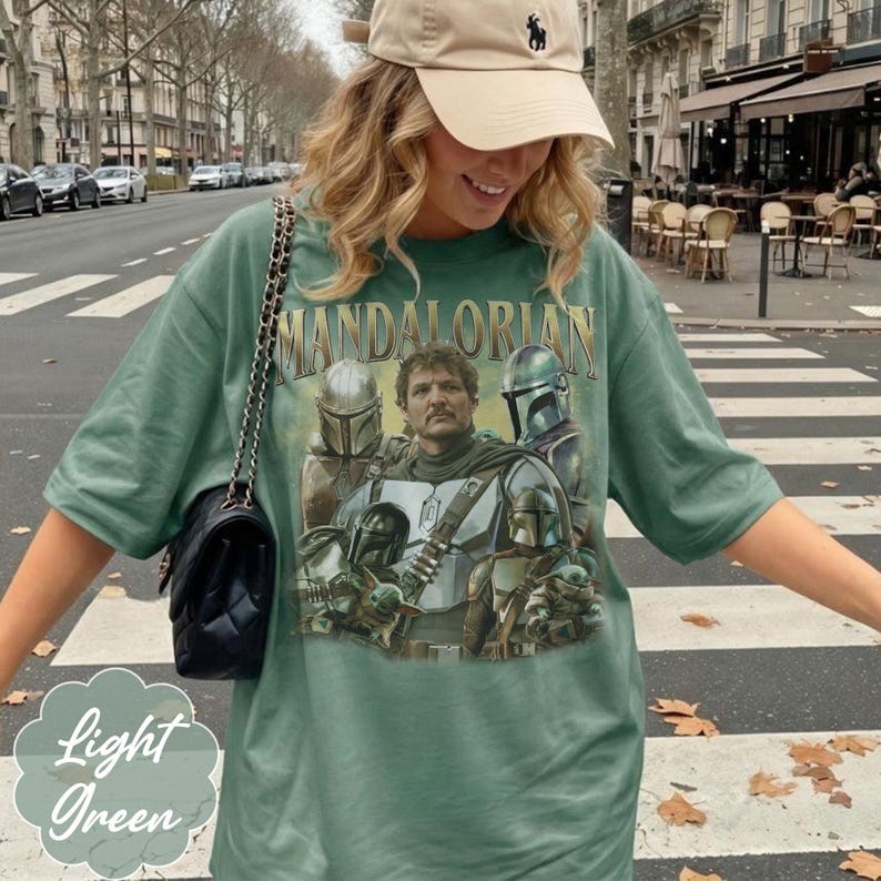 Pu&ograve; includere: Una t-shirt a maniche corte color verde salvia con una stampa grafica dei personaggi di The Mandalorian e il testo "MANDALORIAN" in un carattere dorato invecchiato. Il design include diverse figure corazzate e una piccola creatura.