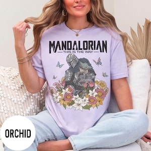 Puede incluir: Camiseta lila con un dise&ntilde;o de The Mandalorian y Grogu rodeados de flores y mariposas. El texto "MANDALORIAN - THIS IS THE WAY -" est&aacute; impreso en negro. La camiseta es de color lila claro.