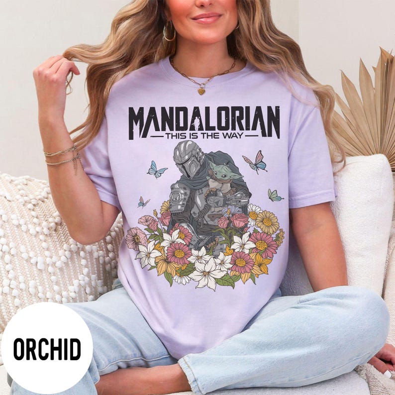 Puede incluir: Camiseta lila con un dise&ntilde;o de The Mandalorian y Grogu rodeados de flores y mariposas. El texto "MANDALORIAN - THIS IS THE WAY -" est&aacute; impreso en negro. La camiseta es de color lila claro.