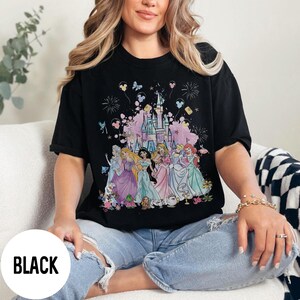 May include: Black t-shirt featuring a colorful graphic of Disney princesses in front of a castle. The design includes fireworks, butterflies, and Mickey Mouse balloons. The word "BLACK" is printed on the bottom left.