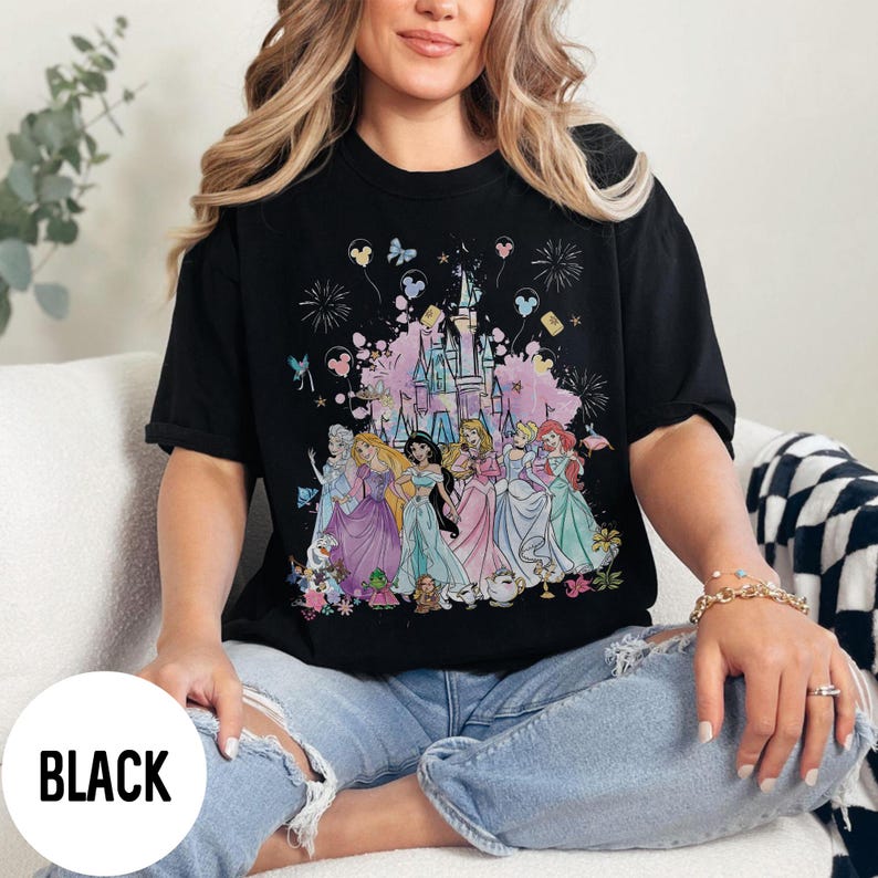May include: Black t-shirt featuring a colorful graphic of Disney princesses in front of a castle. The design includes fireworks, butterflies, and Mickey Mouse balloons. The word "BLACK" is printed on the bottom left.