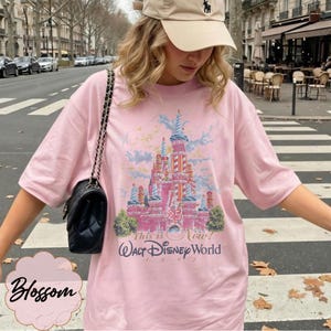 K&ouml;nnte beinhalten: Ein hellrosa T-Shirt mit einer Grafik eines Schlosses in Blau- und Rosat&ouml;nen, dem Text 25, This is Now! und Walt Disney World 1997. Das T-Shirt hat kurze &Auml;rmel und einen Rundhalsausschnitt, kombiniert mit einer schwarzen Umh&auml;ngetasche mit Kette.