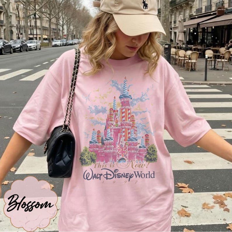 K&ouml;nnte beinhalten: Ein hellrosa T-Shirt mit einer Grafik eines Schlosses in Blau- und Rosat&ouml;nen, dem Text 25, This is Now! und Walt Disney World 1997. Das T-Shirt hat kurze &Auml;rmel und einen Rundhalsausschnitt, kombiniert mit einer schwarzen Umh&auml;ngetasche mit Kette.