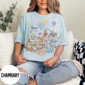 以下が含まれることがあります： 様々な動物のキャラクター、風船、ケーキが描かれた、カラフルな誕生日パーティーのグラフィックが特徴のライトブルーのTシャツ。「Name」という文字と数字の「3」がプリントされています。