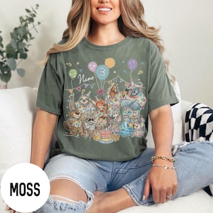 以下が含まれることがあります： モスグリーンのTシャツ。誕生日を祝う動物たちのカラフルな漫画イラストが描かれています。デザインには風船、ケーキ、数字の3が含まれています。「Name」の文字もTシャツに表示されています。