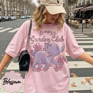 Op de afbeelding: Lichtroze T-shirt met de tekst "Lumpy's Garden Club" en een paarse en roze olifantenillustratie. Het shirt heeft ook het woord "Blossom".