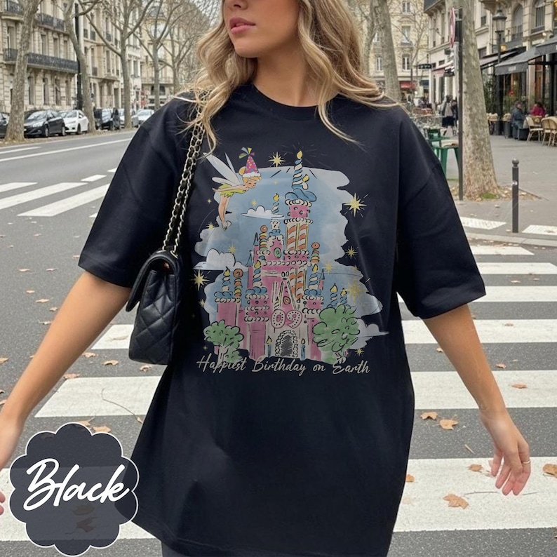 Puede incluir: Camiseta negra con una colorida ilustraci&oacute;n de un castillo de cuento de hadas, un hada y el texto "Happiest Birthday on Earth". El dise&ntilde;o incluye detalles en rosa, azul y amarillo. La camiseta es de estilo casual y oversize.