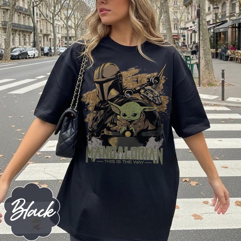 Może przedstawiać: Czarny t-shirt z grafiką przedstawiającą The Mandalorian i Grogu, z napisem "MANDALORIAN - THIS IS THE WAY" w kolorze zielonym i złotym. Koszulka ma kr&oacute;tkie rękawy i luźny kr&oacute;j.