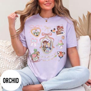 May include: A light purple t-shirt featuring various cartoon character illustrations. The shirt has a round neck and short sleeves. The word "ORCHID" is printed on the bottom left of the shirt.