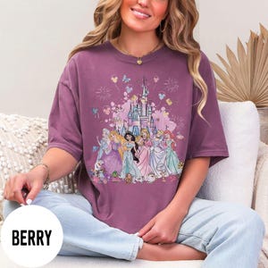 May include: Berry-colored t-shirt featuring a graphic of a castle and several princesses. The design includes fireworks, butterflies, and balloons. The shirt is a short-sleeved, oversized style.