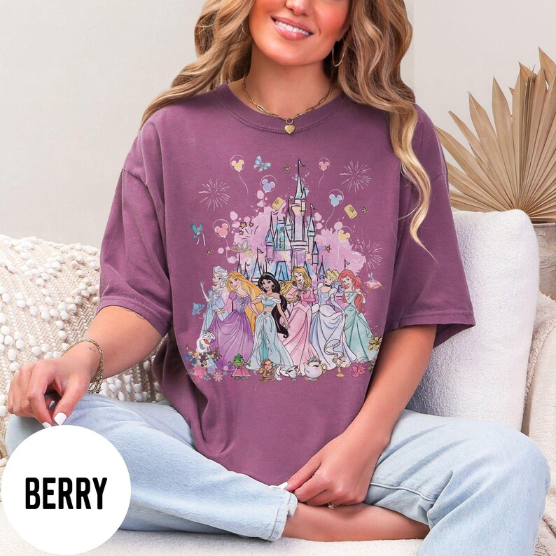 May include: Berry-colored t-shirt featuring a graphic of a castle and several princesses. The design includes fireworks, butterflies, and balloons. The shirt is a short-sleeved, oversized style.