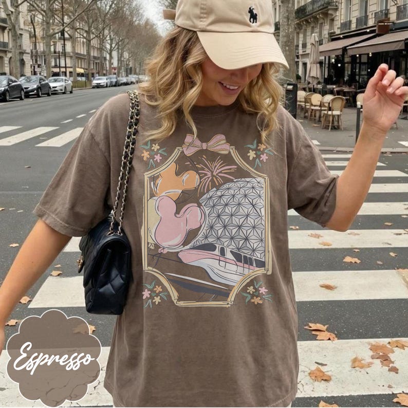 May include: Espresso-colored t-shirt featuring a framed graphic of a theme park scene with Mickey Mouse, a geometric sphere, and a monorail. The shirt has a relaxed fit and short sleeves. A beige baseball cap is worn.