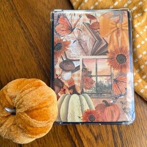 Puede incluir: Funda protectora transparente con un diseño de temática otoñal que presenta calabazas, girasoles, mariposas y un libro abierto. Una calabaza naranja de felpa está al lado de la funda. La funda tiene un borde transparente.