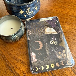 Puede incluir: Una funda decorativa para tableta con un diseño celestial que presenta un sol, una luna, estrellas e imágenes de cartas del tarot. La funda es negra con detalles dorados y blancos. Un cuenco de cerámica azul y una vela también están en la imagen.