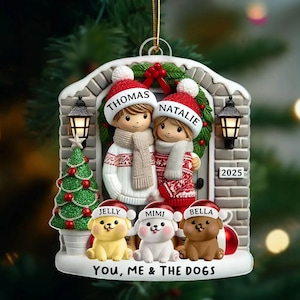 May include: A Christmas ornament featuring a couple and three dogs, all wearing Santa hats. The ornament has the names "Thomas" and "Natalie" on the hats of the couple, and "Jelly", "Mimi", and "Bella" on the dogs' hats. The text "YOU, ME & THE DOGS" is at the bottom.