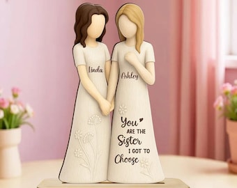 May include: A decorative wooden figurine featuring two figures with brown and blonde hair wearing cream-colored dresses. The figure on the left is labeled Linda, and the right is labeled Ashley with the text, You are the sister I got to choose.