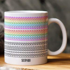 May include: White ceramic mug featuring a colorful rainbow pattern of stitched designs. The mug has a white handle and the name "SOPHIA" printed in black at the bottom. The mug is sitting on a wooden surface.