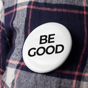 May include: A white, oval-shaped button with the words "BE GOOD" in bold, black letters. The button is pinned to a navy blue and red plaid shirt.