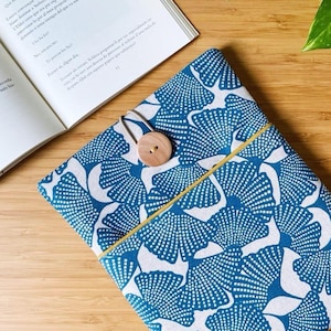 Puede incluir: Una funda de libro con estampado azul y blanco con cierre de botón de madera. La funda presenta un diseño repetitivo en forma de abanico. Un libro abierto descansa junto a la funda sobre una superficie de madera.