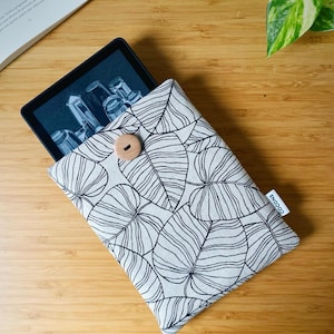 Può includere: Una custodia per e-reader con un motivo a foglie bianche e nere, chiusa con un bottone di legno. La custodia contiene un e-reader nero che mostra un'immagine. Una piccola etichetta bianca con la scritta "cocons" è attaccata.