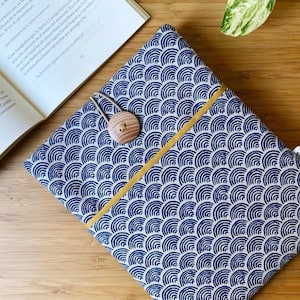 Puede incluir: Una funda de libro con estampado azul y blanco con cierre de botón de madera. La funda presenta un diseño de olas repetidas y una raya amarilla. Un libro abierto es visible en el fondo.