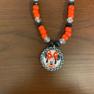May include: A necklace with orange, black, and clear beads, and a Minnie Mouse pendant. The pendant shows Minnie Mouse with a red bow and a polka dot border. The necklace is a fun accessory.