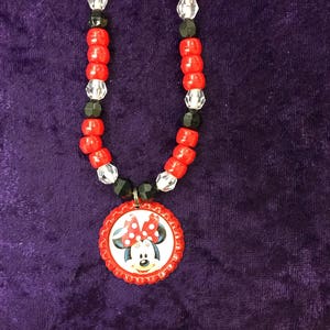 May include: A red beaded necklace with a Minnie Mouse pendant. The necklace features red, black, and clear beads. The pendant is a red bottle cap with a Minnie Mouse graphic.