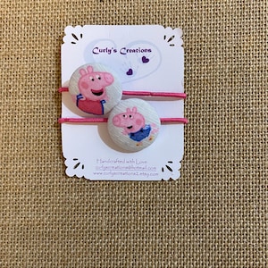May include: Two pink hair ties with fabric buttons featuring Peppa Pig characters. The buttons are white with pink and blue details. The hair ties are on a white card with the text "Curly's Creations" and contact information.