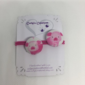 May include: Two pink pig hair ties with white polka dots. The hair ties are on a white card with the text "Curly's Creations" and contact information.