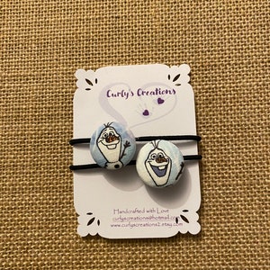 May include: Two blue and white fabric hair ties with Olaf from Frozen printed on them. The hair ties are on a white card with the text "Curly's Creations" and "Handcrafted with Love".