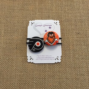 May include: Two fabric hair scrunchies with a black and orange hockey team logo and a cartoon character with an orange beard. The scrunchies are on a white card with the text "Curly's Creations" and "Handcrafted with Love".