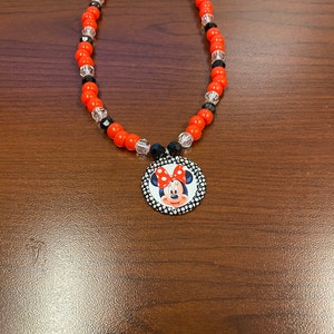 May include: A necklace with red, black, and clear beads, and a Minnie Mouse pendant. The pendant is circular, with a black and white polka dot border and a Minnie Mouse graphic. The necklace is a children's accessory.