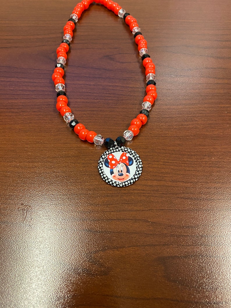May include: A necklace with red, black, and clear beads, and a Minnie Mouse pendant. The pendant is circular, with a black and white polka dot border and a Minnie Mouse graphic. The necklace is a children's accessory.