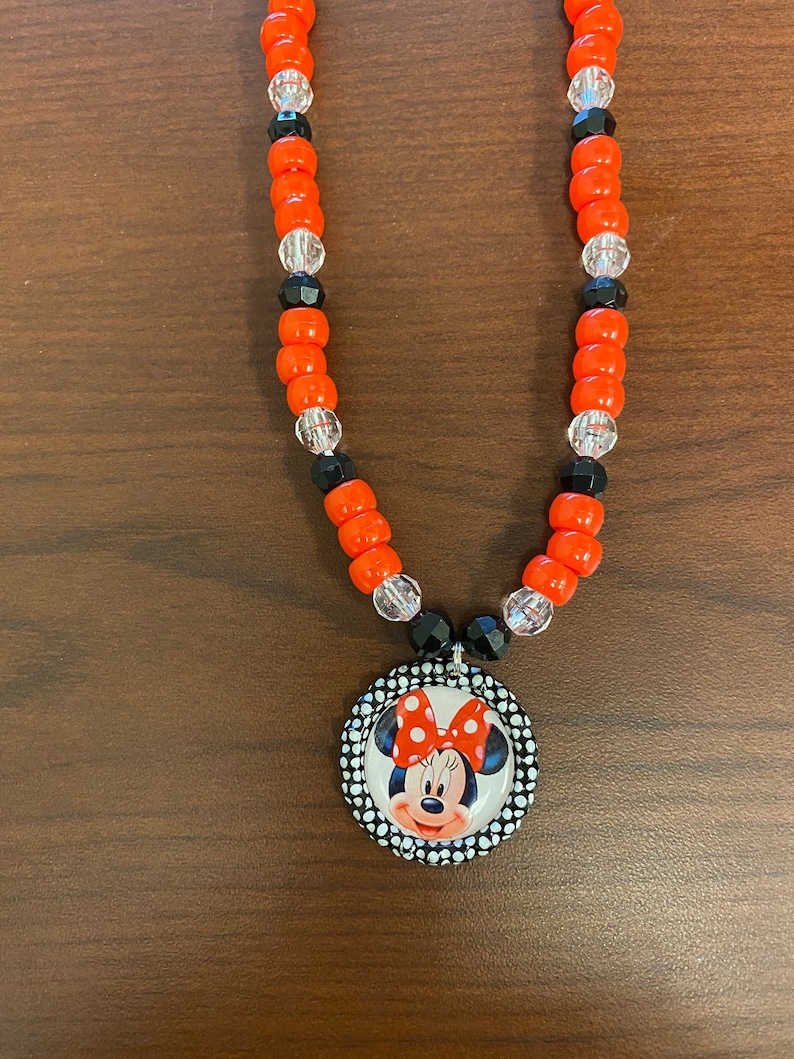 May include: A necklace with orange, black, and clear beads, and a Minnie Mouse pendant. The pendant shows Minnie Mouse with a red bow and a polka dot border. The necklace is a fun accessory.