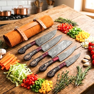 Puede incluir: Un juego de cinco cuchillos de cocina con hojas de acero de Damasco y mangos de madera oscura con detalles en rojo, dispuestos junto a un estuche de cuero beige, rodeados de verduras picadas y hierbas frescas sobre una mesa.