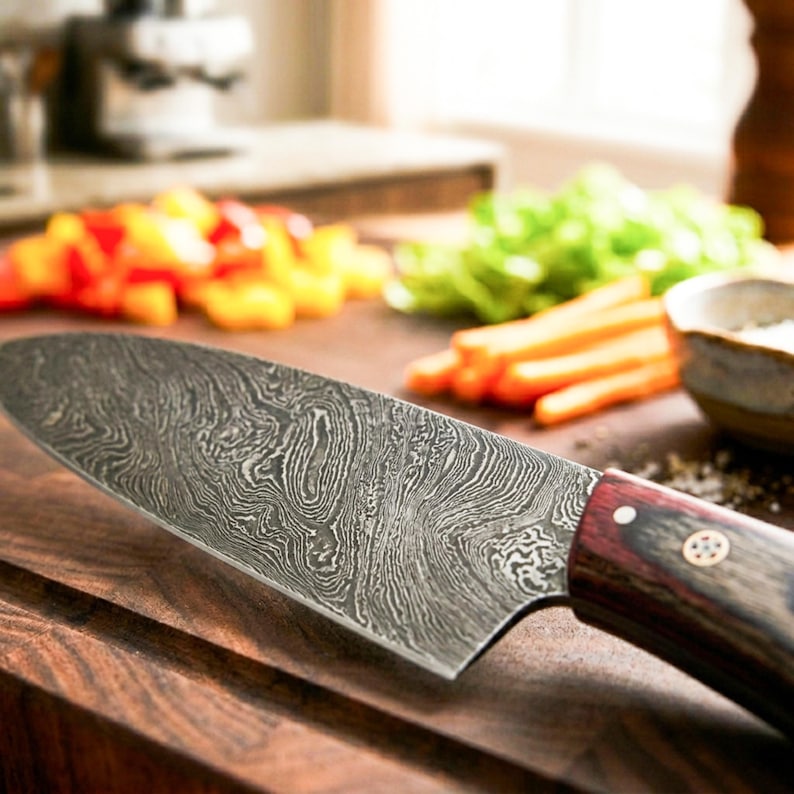 Puede incluir: Un cuchillo de chef con una hoja de acero de Damasco estampada y un mango de madera de color marr&oacute;n rojizo oscuro asegurado con dos remaches met&aacute;licos. La hoja presenta una textura met&aacute;lica ondulada y estratificada con un detalle.