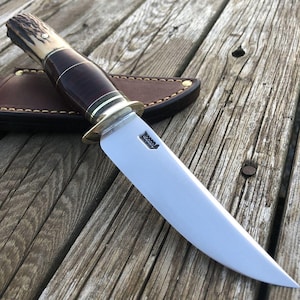 Puede incluir: Un cuchillo de caza con una hoja de acero pulido y un mango marrón oscuro con detalles en latón. El mango tiene una sección de material de color claro cerca de la parte superior. Una funda de cuero marrón es visible en el fondo.