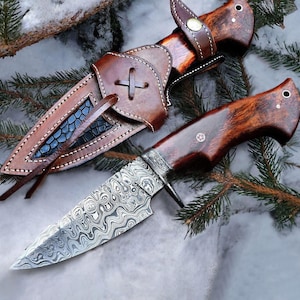 Puede incluir: Un cuchillo de caza artesanal con una hoja de acero de Damasco y un mango de madera. El cuchillo se combina con una funda de cuero marrón. La funda presenta un diseño en forma de cruz y costuras decorativas. El cuchillo y la funda se colocan sobre un fondo nevado.