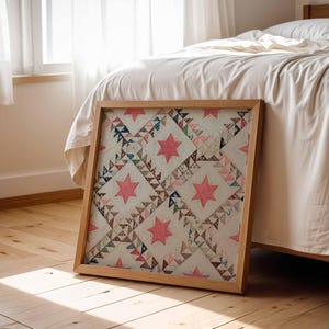 May include: A framed quilt with a wooden frame, featuring a repeating pattern of pink stars and geometric shapes in various colors. The quilt is leaning against a bed with white bedding, on a wooden floor.