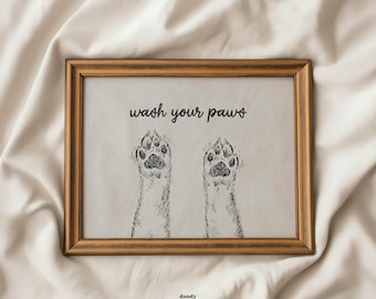 May include: A framed print with a light brown wooden frame. The print features two sketched dog paws with the text "wash your paws" above them. The words "dandy prints" are at the bottom of the print.