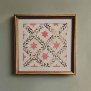 May include: Framed quilt art featuring a star pattern in pink and various muted colors. The quilt is framed in a gold-colored wooden frame. The quilt design includes pink stars and geometric shapes.
