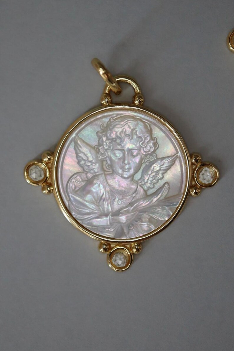 May include: Gold pendant featuring a carved mother-of-pearl cameo of an angel with wings. The pendant has a round frame with four diamond accents and a loop for a chain. The angel is depicted in a classical style.