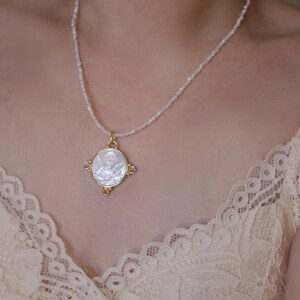 May include: A delicate necklace featuring a gold pendant with a white, circular inlay. The pendant is adorned with a detailed design and hangs from a beaded chain. The necklace is displayed against a soft, neutral background.
