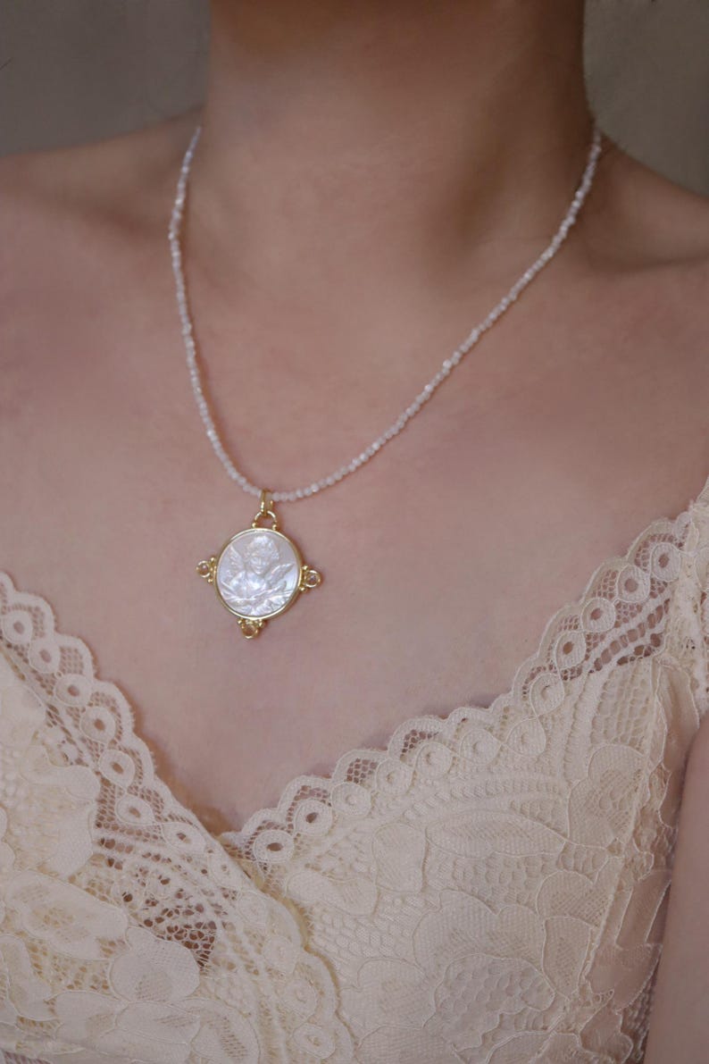 May include: A delicate necklace featuring a gold pendant with a white, circular inlay. The pendant is adorned with a detailed design and hangs from a beaded chain. The necklace is displayed against a soft, neutral background.