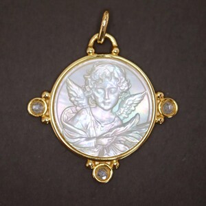 May include: A gold pendant featuring a carved mother-of-pearl cameo of an angel with wings. The pendant has a circular design with a gold frame and is adorned with three clear gemstones. The pendant is suspended from a gold bail.