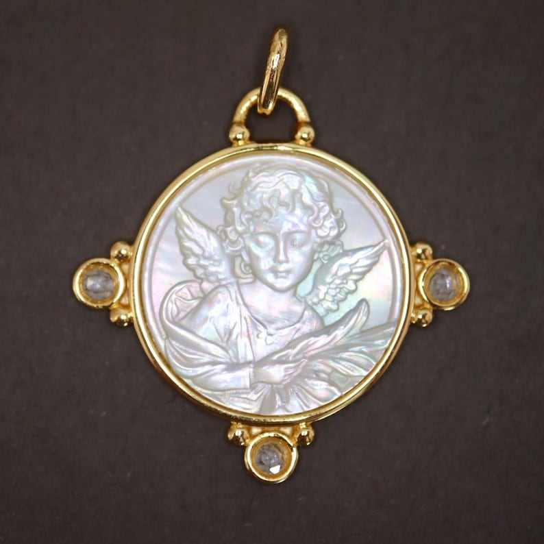May include: A gold pendant featuring a carved mother-of-pearl cameo of an angel with wings. The pendant has a circular design with a gold frame and is adorned with three clear gemstones. The pendant is suspended from a gold bail.