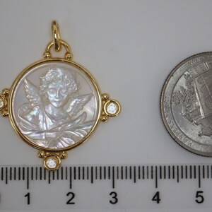 May include: A gold pendant featuring a carved angel design within a circular frame. The pendant has a gold bail and is adorned with small, clear gemstones at each corner. A US quarter is next to the pendant for size comparison.