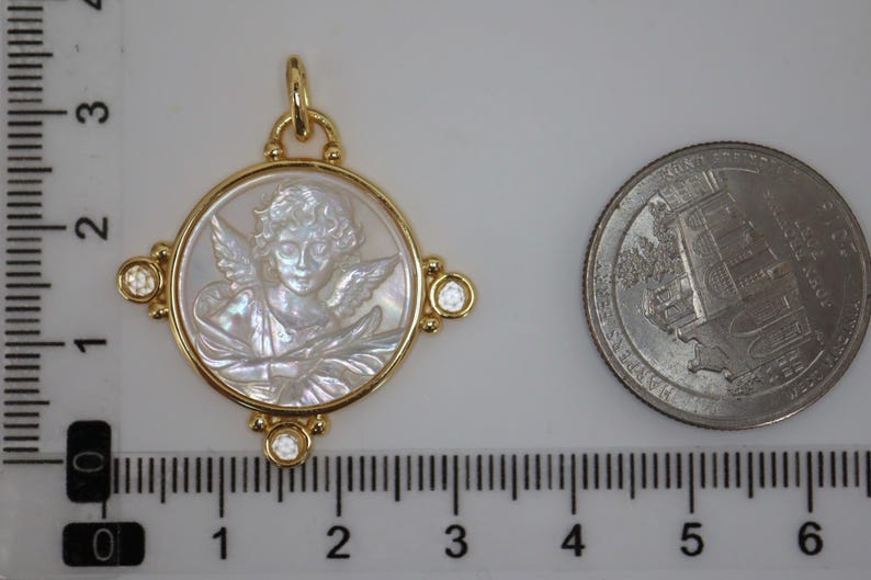 May include: A gold pendant featuring a carved angel design within a circular frame. The pendant has a gold bail and is adorned with small, clear gemstones at each corner. A US quarter is next to the pendant for size comparison.