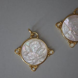 May include: Two gold-toned pendants with a circular design featuring a carved cherub image. The cherub is depicted with wings and curly hair. The pendants have a gold frame with small, clear gemstones at the corners. The background is a solid gray.