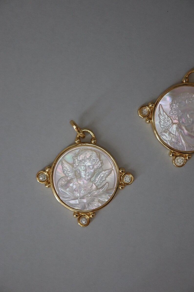 May include: Two gold-toned pendants with a circular design featuring a carved cherub image. The cherub is depicted with wings and curly hair. The pendants have a gold frame with small, clear gemstones at the corners. The background is a solid gray.